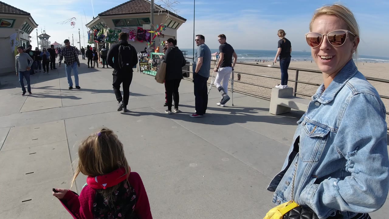 At 0:01 in Huntington Beach, California, the child remains in the lower foreground while the woman in sunglasses fills the right side and pedestrians move near vendor displays by the ocean railing.