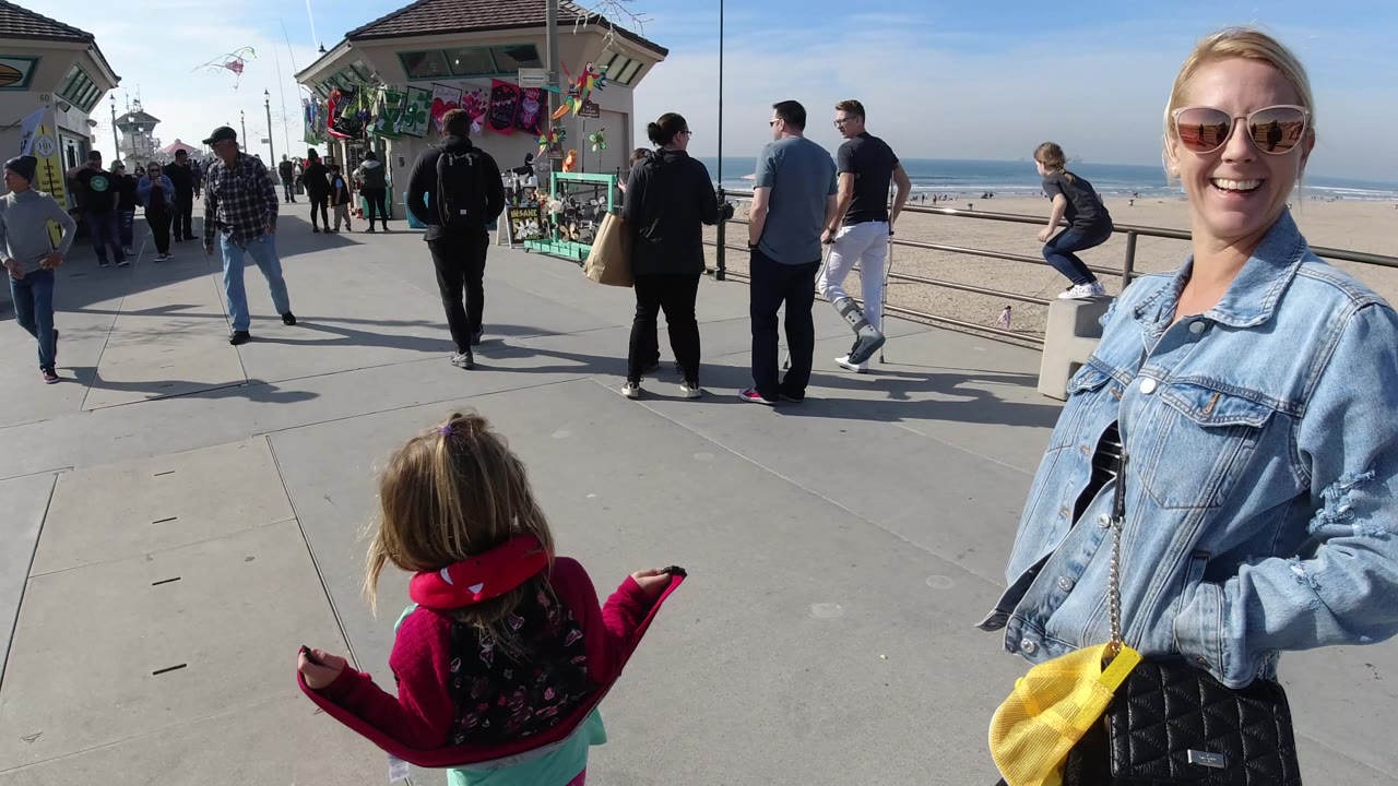 At 0:02 in Huntington Beach, California, the smiling woman is cropped along the right edge while the child walks ahead and several visitors stand near the railings with the beach and surf beyond.