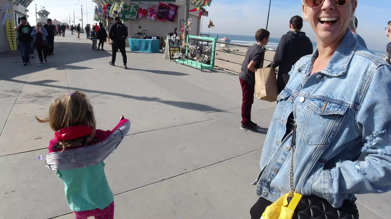 At 0:00 in Huntington Beach, a child carries an inflatable dolphin while a smiling woman in sunglasses stands close to camera on a busy concrete pier walkway.