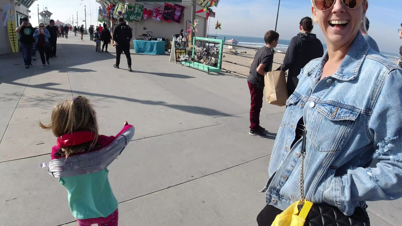 At 0:01 in Huntington Beach, the child remains centered with the inflatable dolphin across their shoulders as pedestrians and colorful vendor displays line the pier walkway.