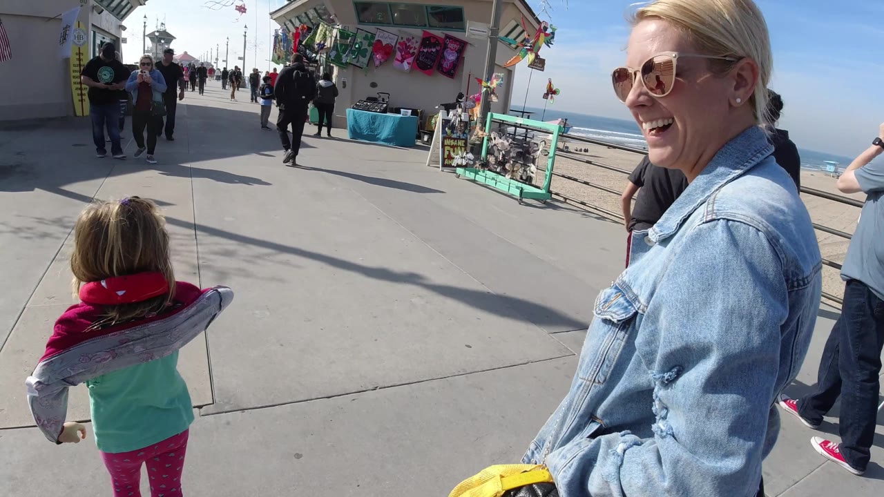 At 0:02 in Huntington Beach, the smiling woman is cropped at the right edge while the child with the inflatable dolphin faces down the pier toward pedestrians and vendor tables.