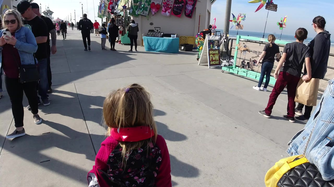 Child in stroller near adults walking past a pier-side vendor table.