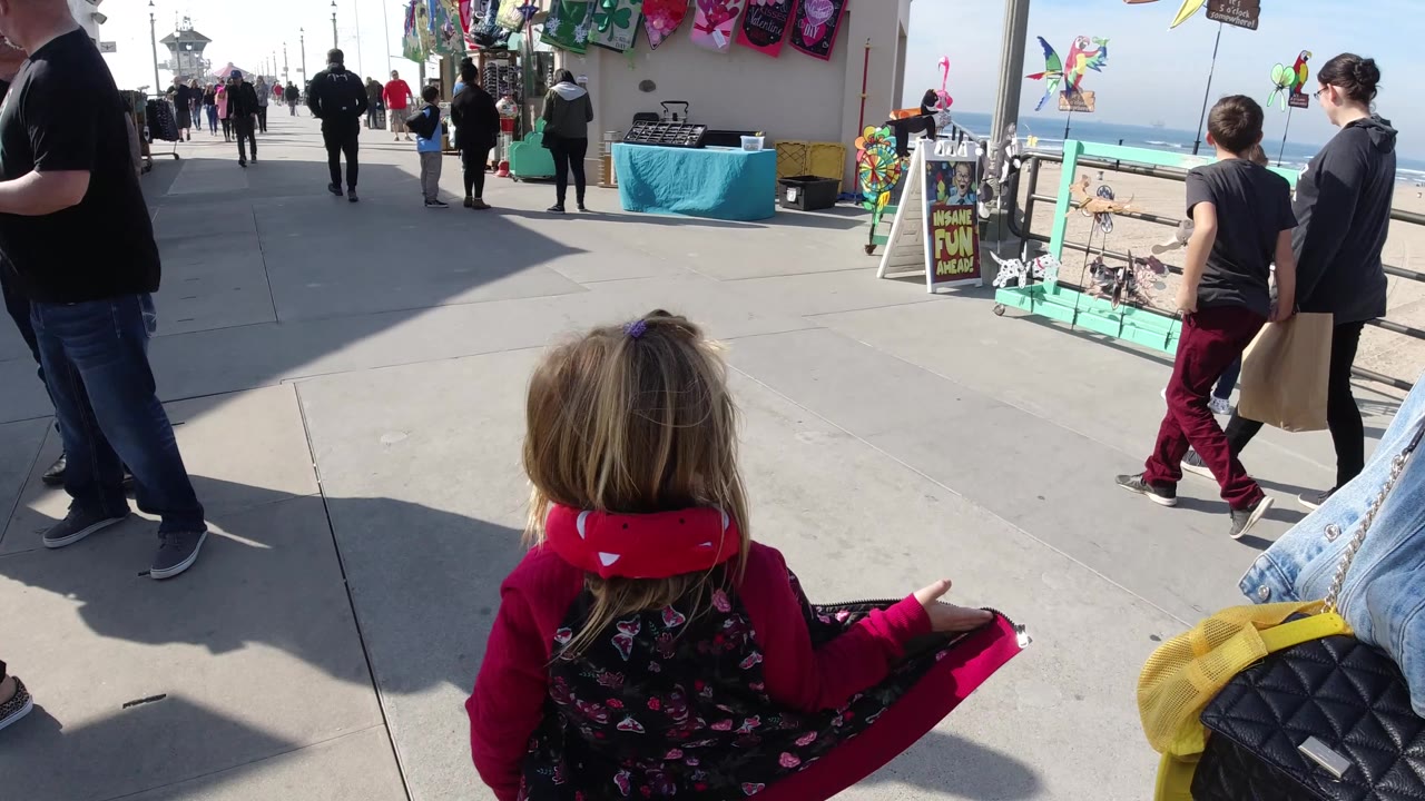 A child sits in the stroller at right of the lower frame while a man stands close by and other pedestrians pass the Huntington Beach pier vendor display.