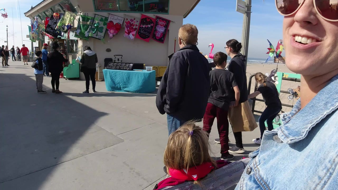 At Huntington Beach, California, a beachside walkway passes a vendor stand with hanging colorful merchandise as adults and children move through bright sun near the ocean railing.