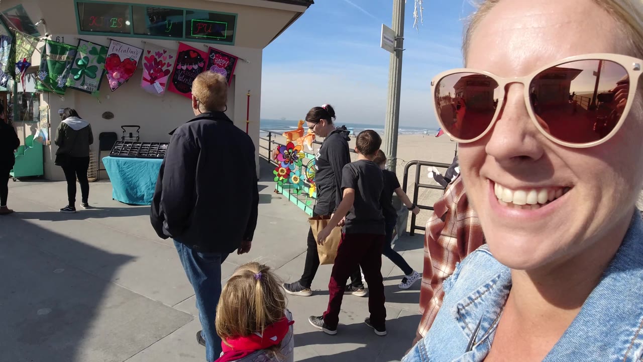 At Huntington Beach, California, a smiling woman in large sunglasses fills the right side of the frame while adults and children walk past a beachside shop and ocean railing.