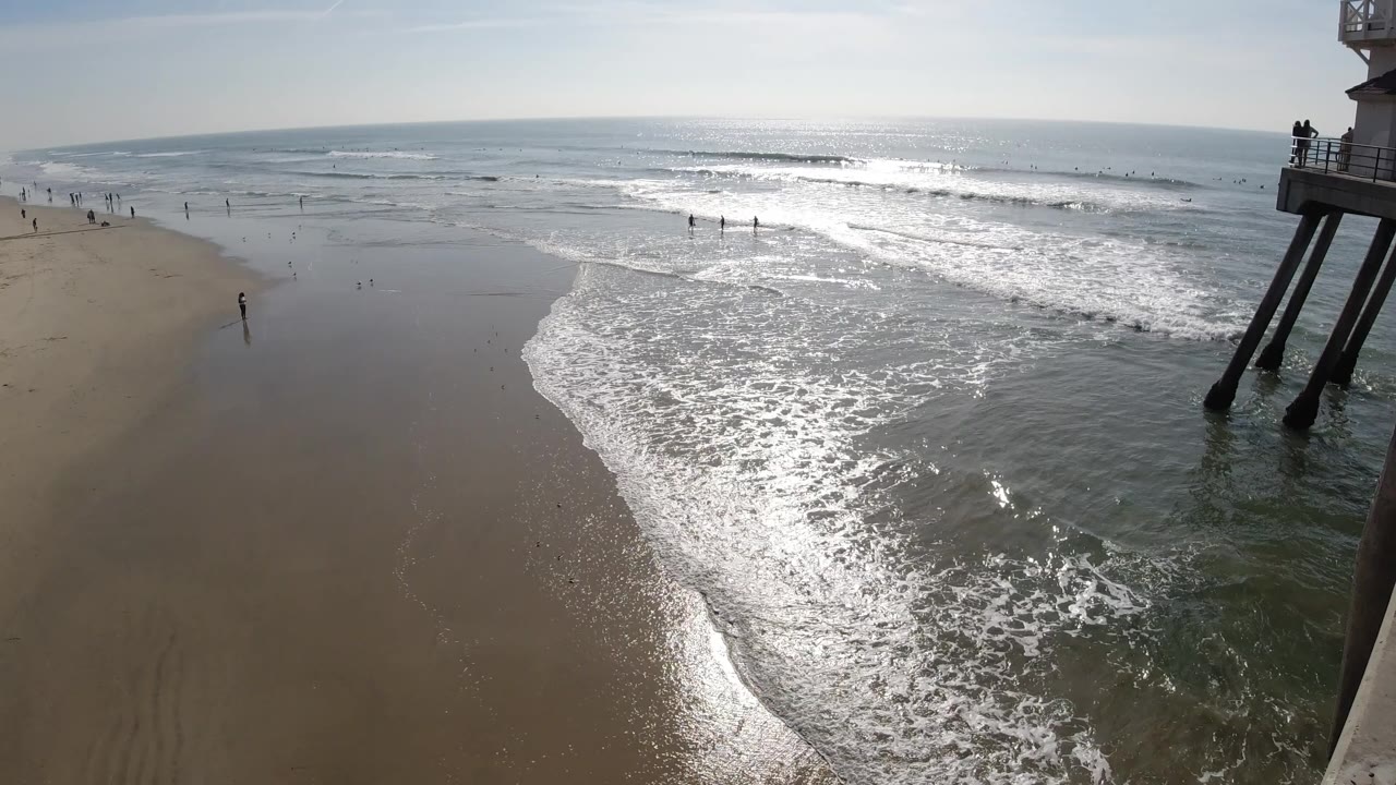 Elevated Huntington Beach view with bright surf curling over wet sand, distant beachgoers, and dark pier supports at the right edge.