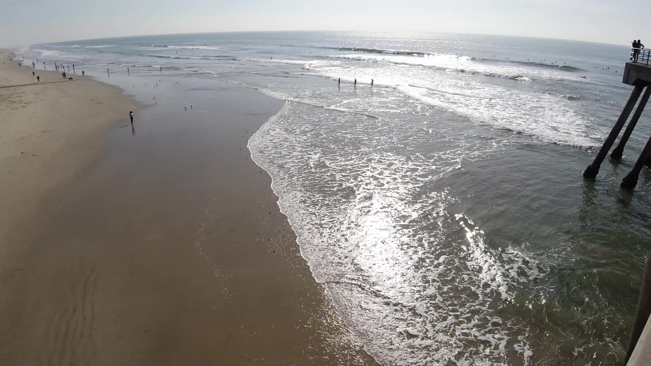 Huntington Beach surf line seen from above, with ocean foam advancing diagonally across sand, distant figures near the water, and cropped pier supports to the right.