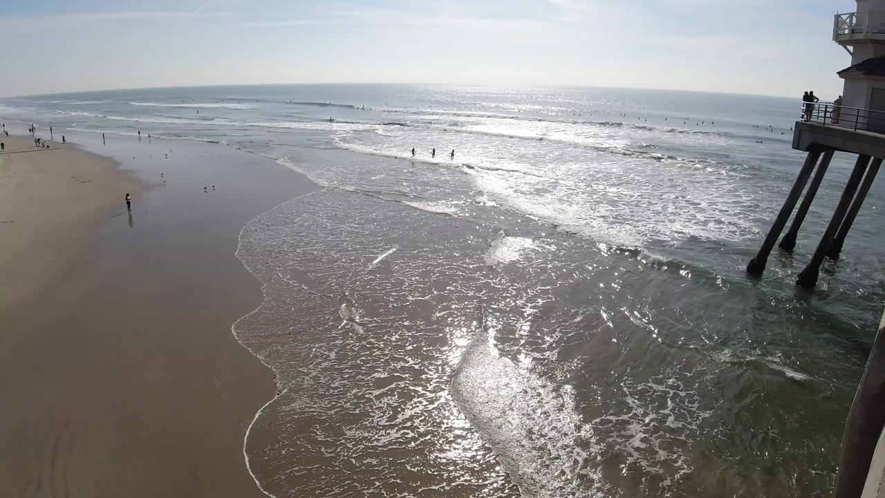 Elevated Huntington Beach view with foamy surf spreading across wet sand, bright sun reflection on the water, distant beachgoers, and pier supports on the right.