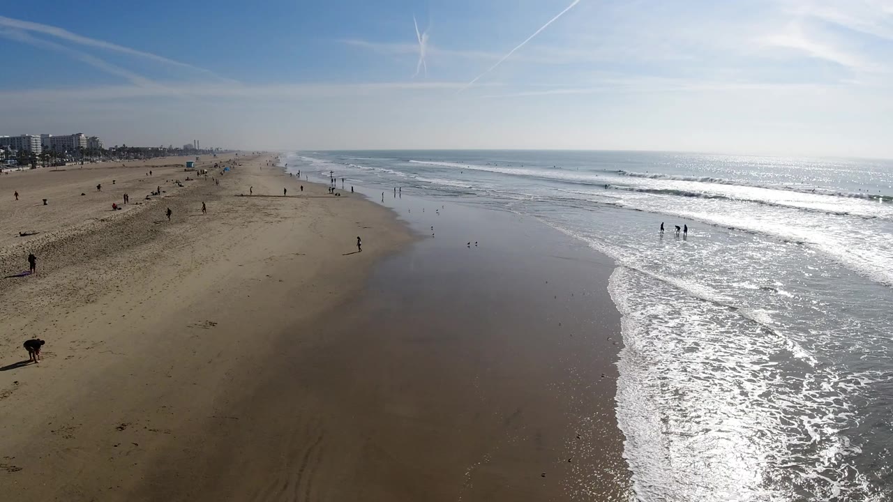 Elevated view over Huntington Beach with wide sand on the left, bright ocean surf on the right, distant buildings near the horizon, and small people visible along the shore.