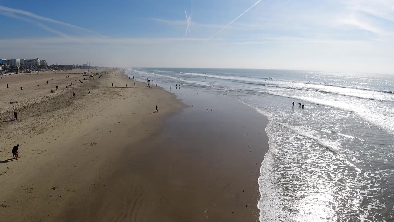 A broad Huntington Beach view shows open sand, a reflective wet shoreline, white wave foam, and several small figures spaced along the beach and shallow water.