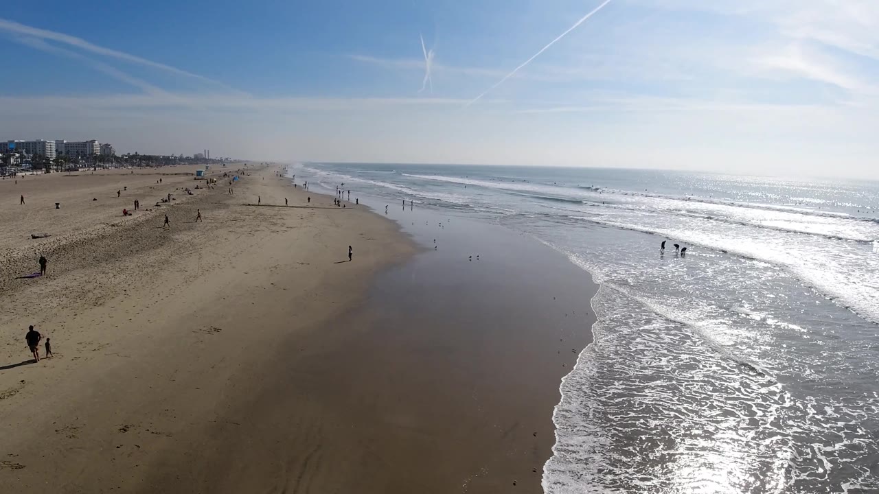 Aerial beach view with surf, reflective water, and scattered people.