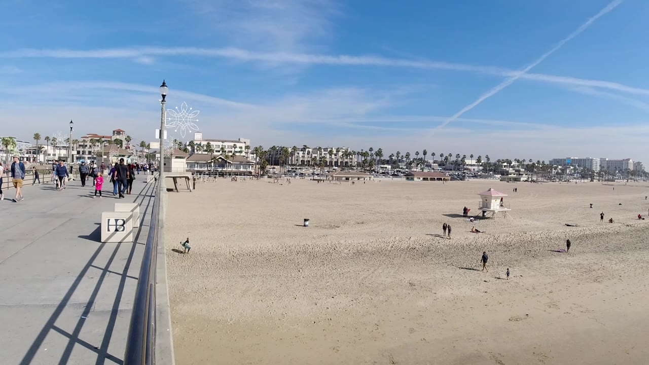 Huntington Beach boardwalk perspective with walkers near the railing, a lifeguard tower on the sand, palm-lined buildings in the background, and pale contrails crossing the blue sky.