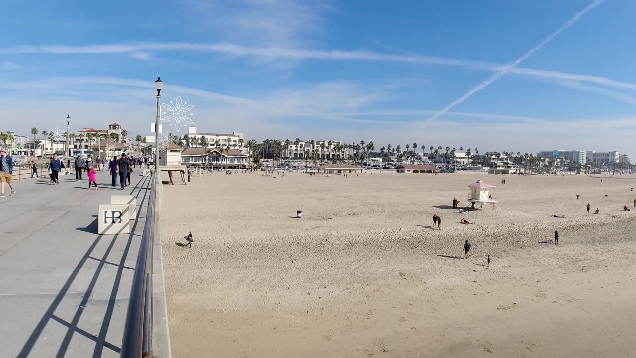 Elevated Huntington Beach frame showing the boardwalk railing near center, people walking on the promenade, scattered figures on the sand, a lifeguard tower, palms, and low coastal buildings.
