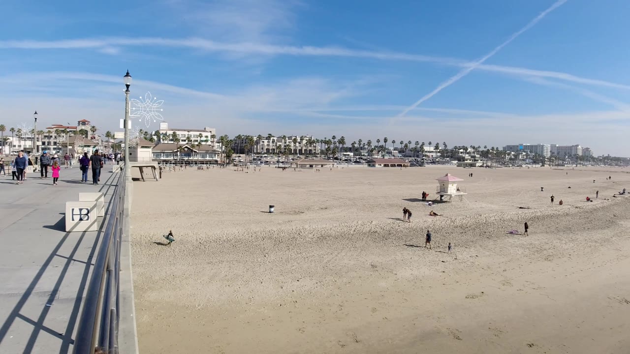 Boardwalk and beach scene with people, railings, open sand, palm trees, and a lifeguard tower.