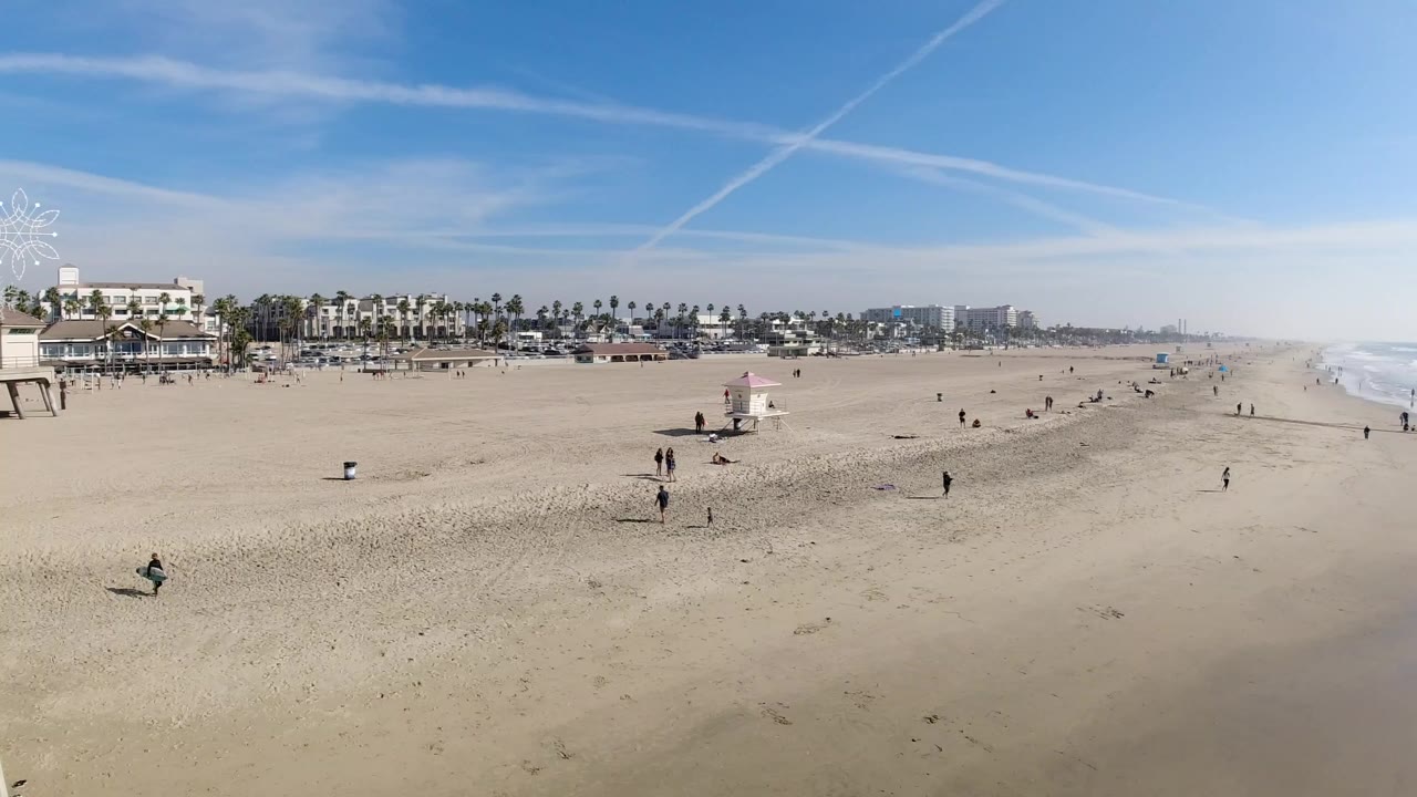Wide elevated view of Huntington Beach, California, with scattered beachgoers on sand, a small lifeguard tower, palm-lined buildings, and surf running along the right side.