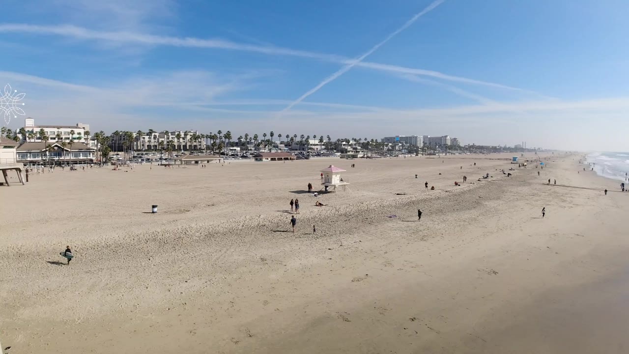 Elevated Huntington Beach view showing the pink-roof lifeguard tower nearer the lower center, small figures on the sand, a line of palms and buildings, and bright water at right.