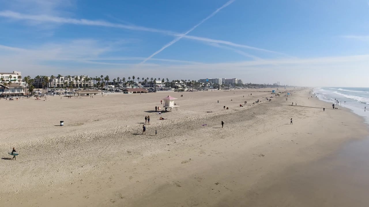 Wide Huntington Beach frame with the shoreline stretching into haze, small beachgoers across the sand, low coastal buildings and palms at left, and white surf lines at right.