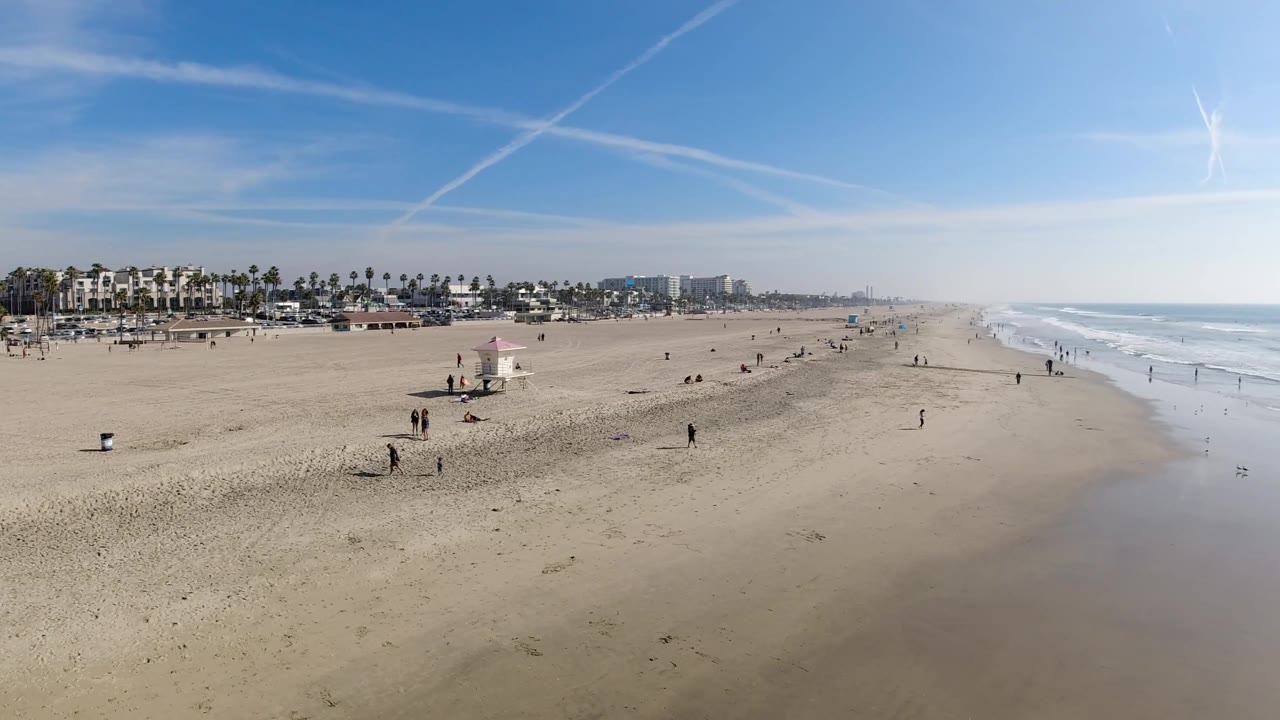 Sunny beach with lifeguard tower, people on sand, palms, and surf to the right.