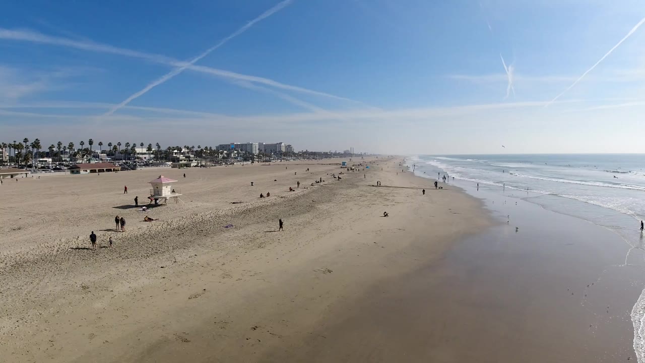 Wide aerial view of Huntington Beach with open sand on the left, shallow waves on the right, small people along the shoreline, and bright blue sky above distant palm-lined buildings.