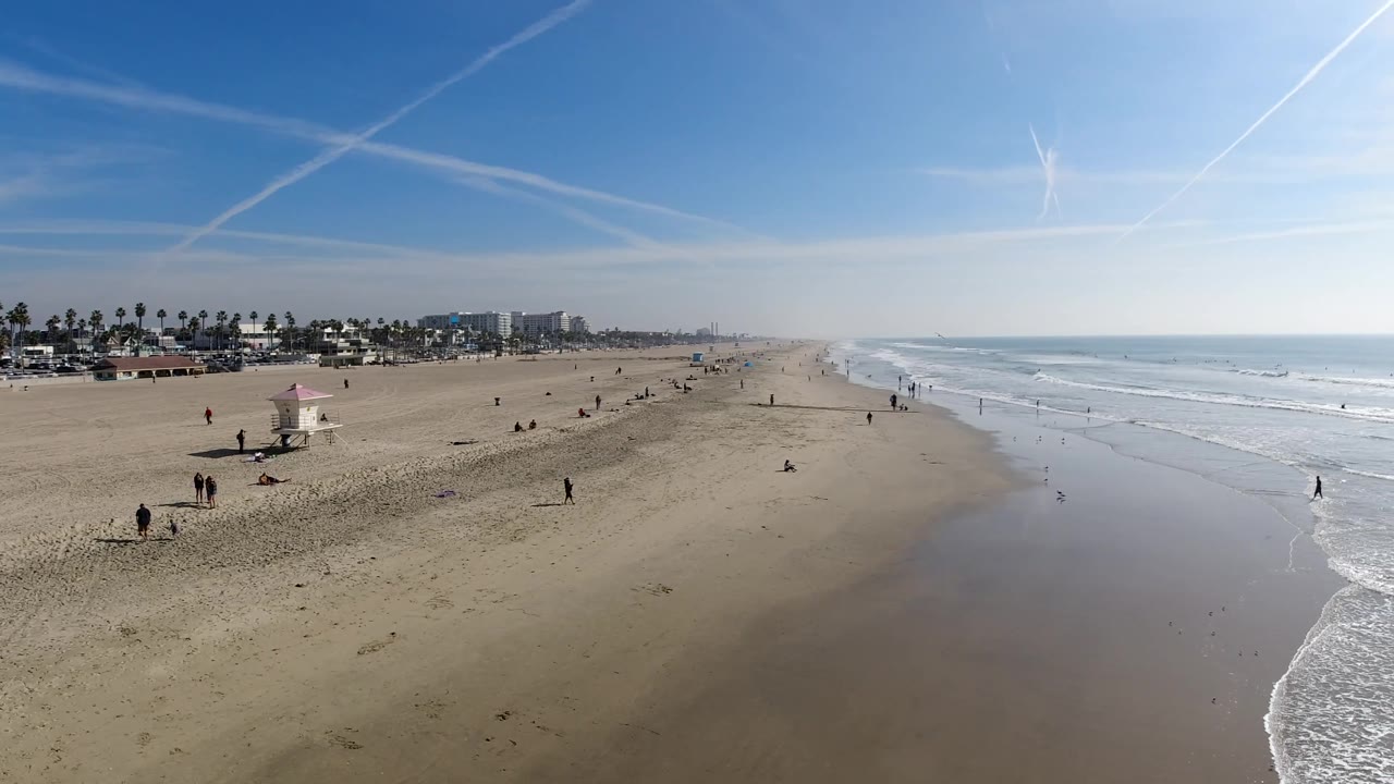 Elevated Huntington Beach frame with the ocean horizon to the right, scattered people near the waterline, open sand in the foreground, and distant beachfront structures at left.