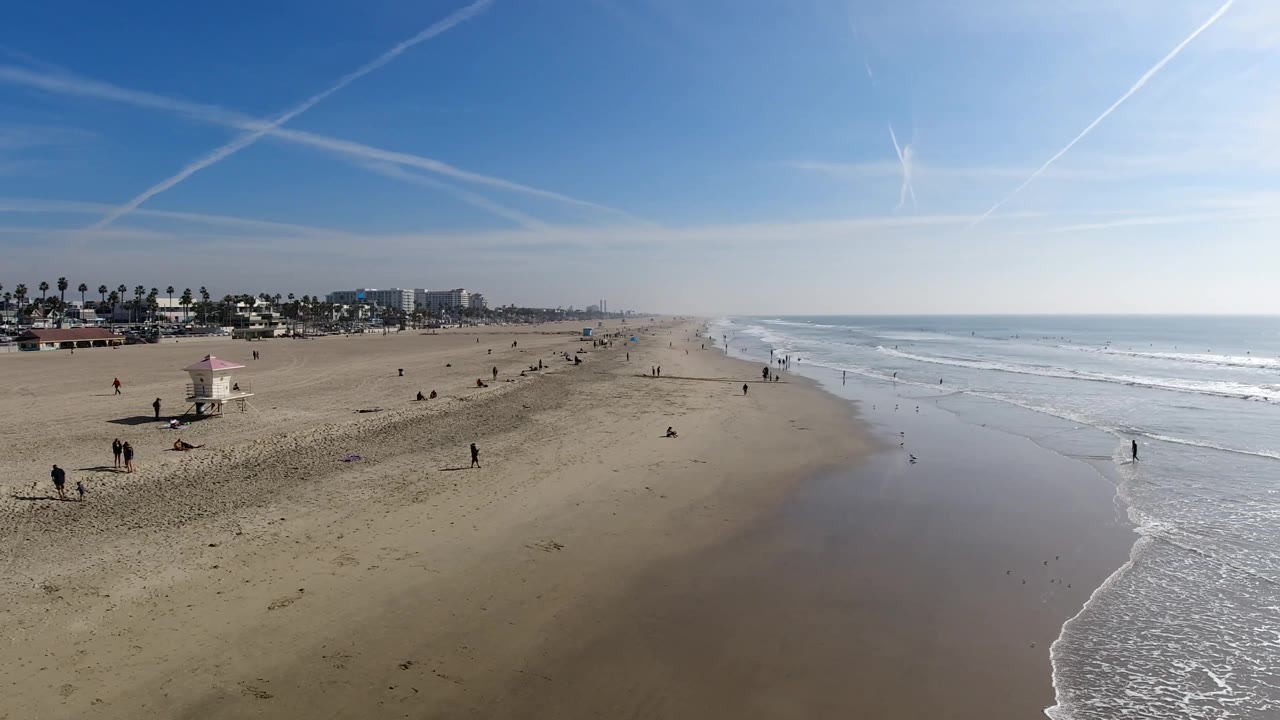 Wide beach aerial with surf approaching the wet sand and people walking below.