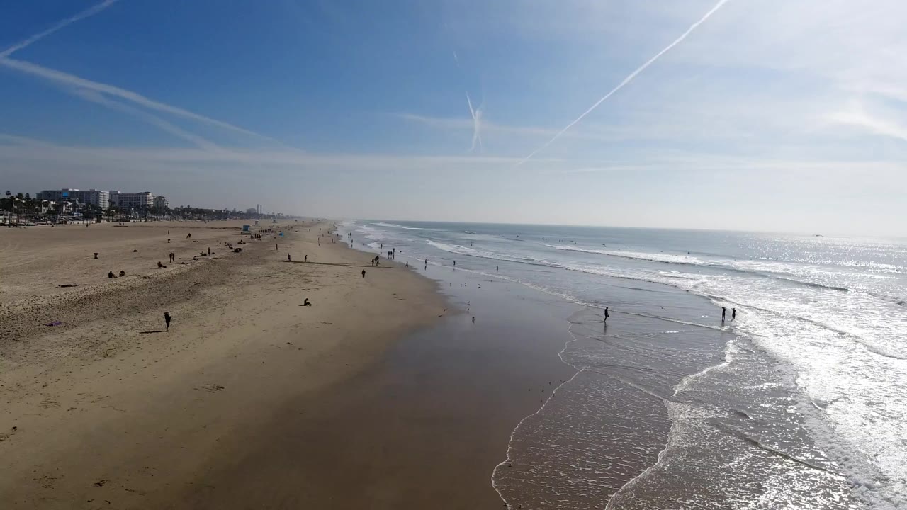 Elevated Huntington Beach, California shoreline view with brighter surf on the right, reflective wet sand through the center, and tiny people dispersed near the water.