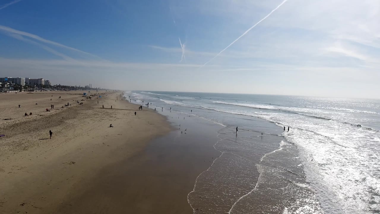Wide Huntington Beach, California coast with open sand on the left, ocean surf on the right, distant buildings, and small figures visible near the waterline.