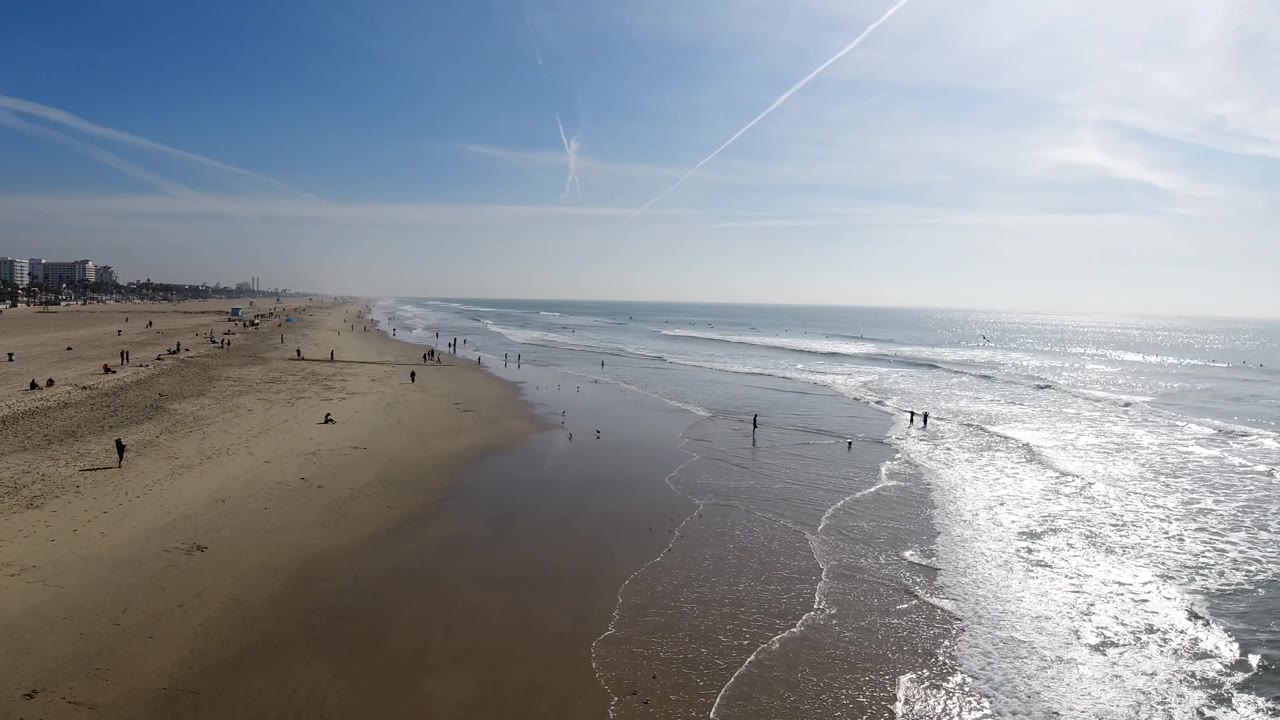 Elevated beach and surf view under bright sky with distant buildings.