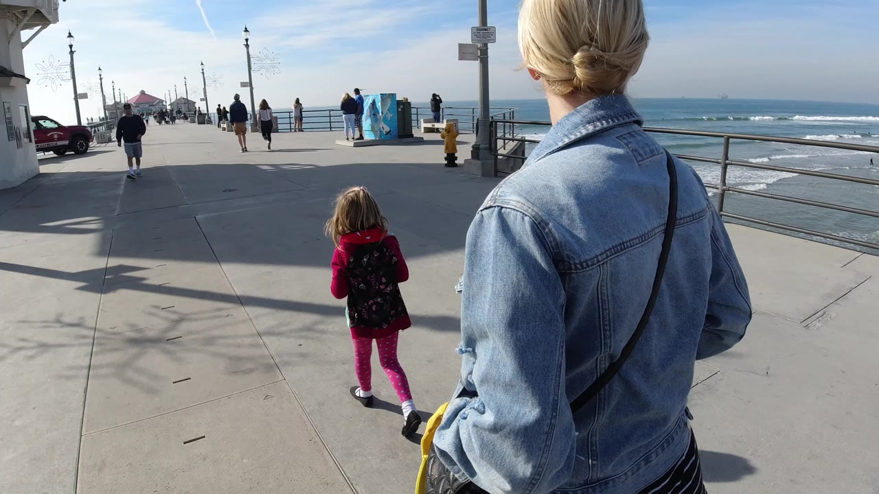 At Huntington Beach, a blonde woman in a denim jacket and a young girl in pink walk away from camera on a sunny pier with ocean railings and pedestrians ahead.