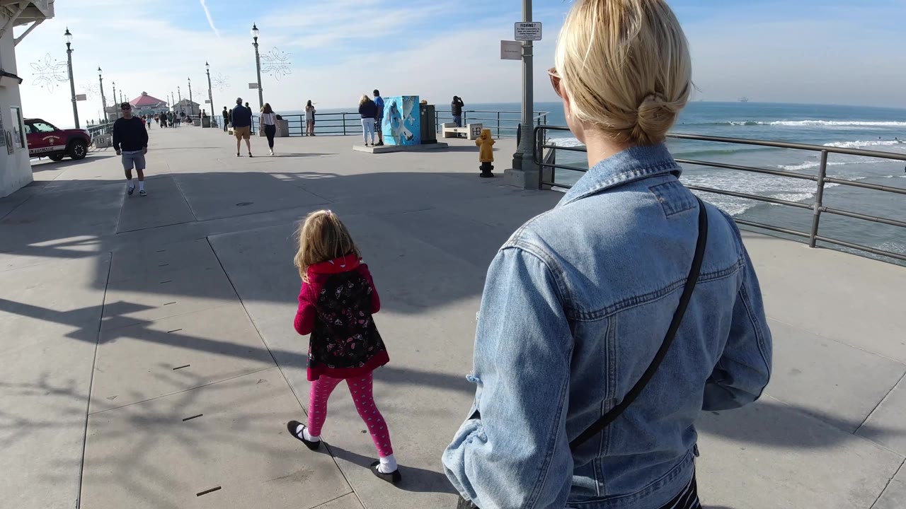 A closer right-side view shows the woman's shoulder and hair in the foreground as the child walks toward the open Huntington Beach pier and ocean horizon.