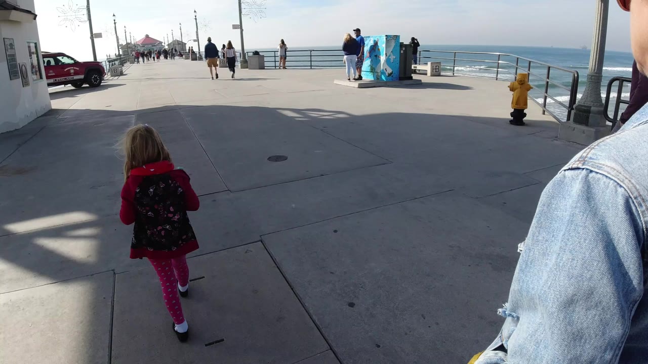A child in a red top and backpack walks ahead on a Huntington Beach concrete promenade with ocean railings, distant pedestrians, a red vehicle, and bright afternoon shadows.