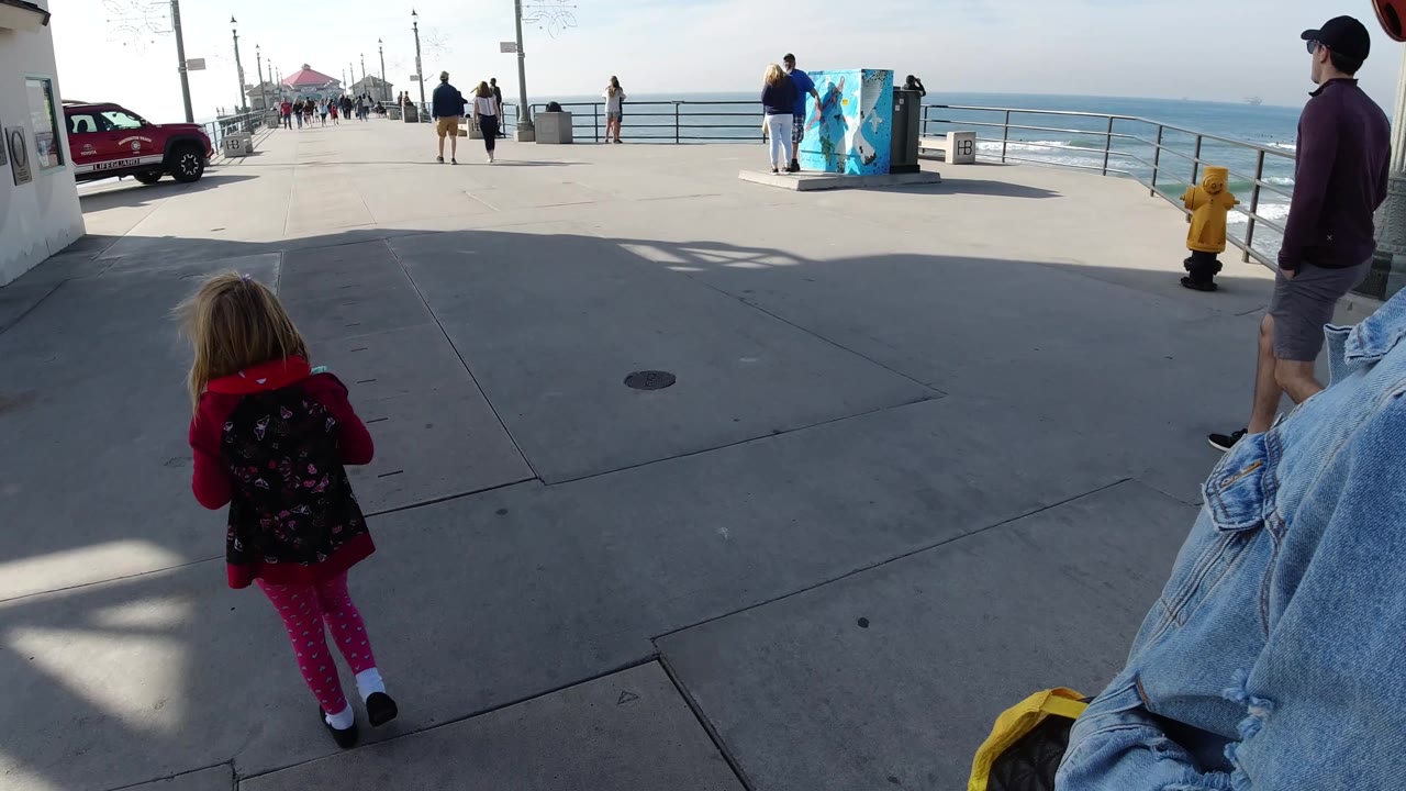 A man in a cap appears at the right edge while the child walks toward the Huntington Beach railings, with the ocean, utility box artwork, and long shadows across the pavement.