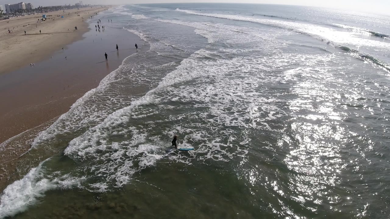 High aerial view over Huntington Beach surf, with foamy water in the foreground, wet sand along the left edge, sun glare on the ocean, and distant people on the beach.