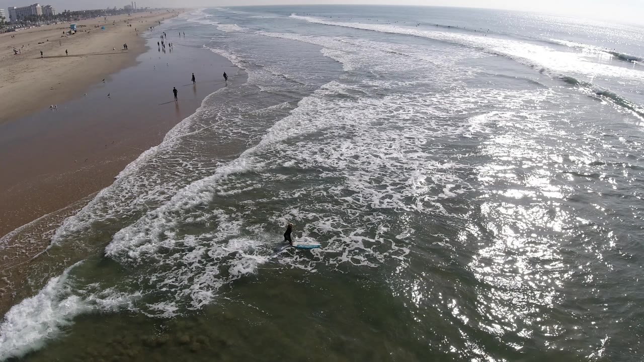 Aerial Huntington Beach frame with a diagonal wave line moving across shallow water, bright reflection on the right, and small figures scattered along the wet sand.