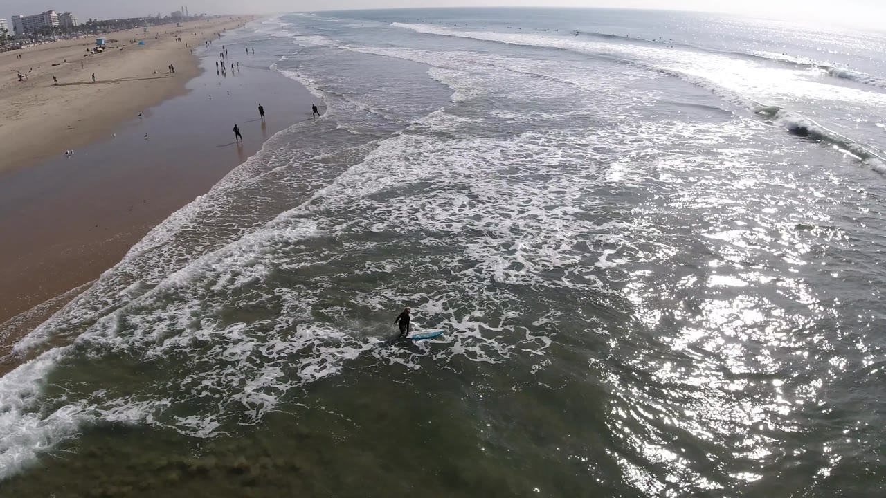 Huntington Beach surf fills the frame, with a small board rider near the lower center, bright ocean glare to the right, and distant walkers along the left shoreline.