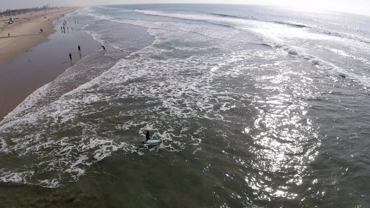 Sunlit surf and wet sand seen from above with distant people near the waterline.