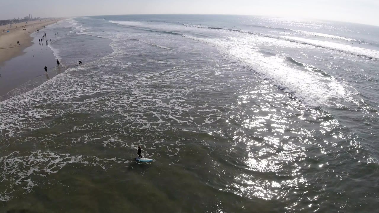 Huntington Beach shoreline seen from above with the surf line stretching across the frame, beachgoers on the sand, and a blue board near the lower center.