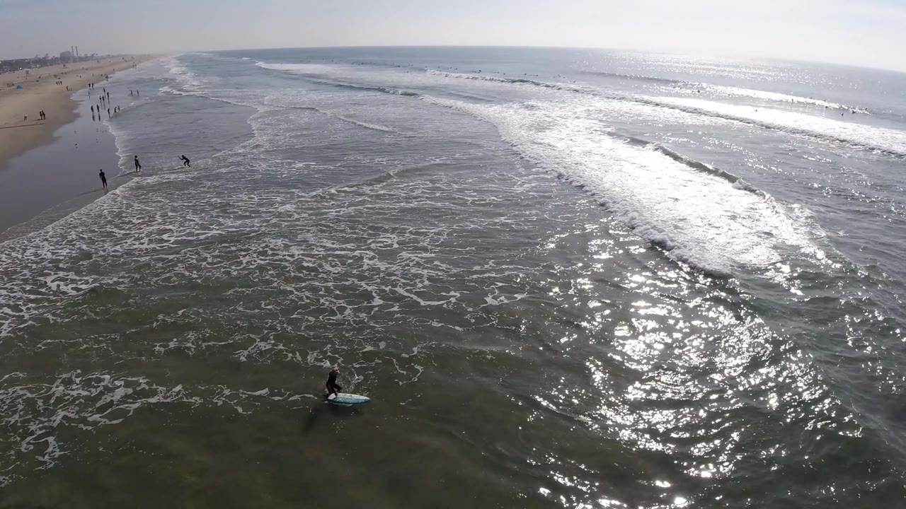 Aerial Huntington Beach view with a broad sheet of foamy water, bright reflections on the right, people along the beach, and a board rider near the lower right edge.