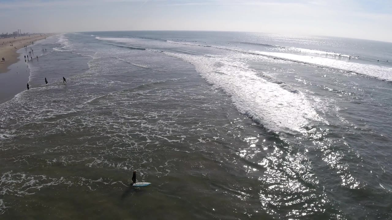 Aerial shoreline scene with rolling surf and a person on a blue board near the bottom.