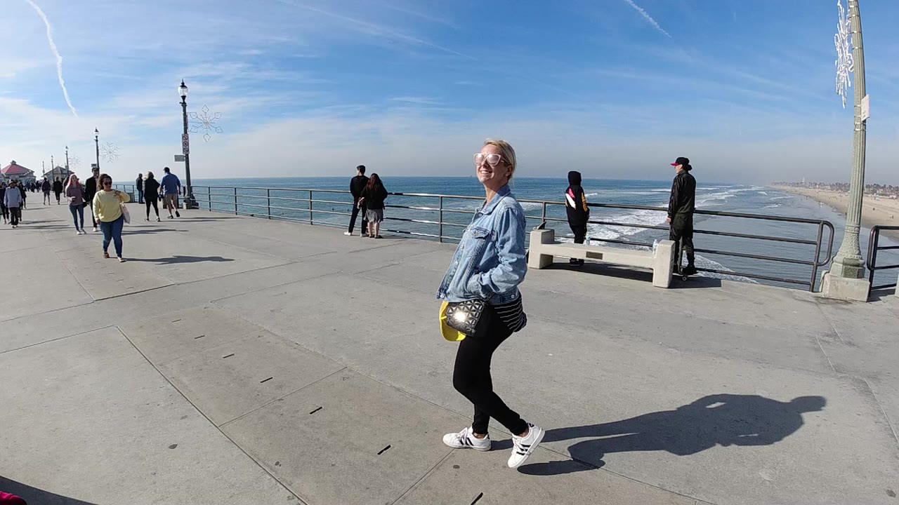 At 0:00, a woman in sunglasses and a denim jacket walks across a Huntington Beach pier as ocean waves, railings, benches, lampposts, and several pedestrians fill the background.