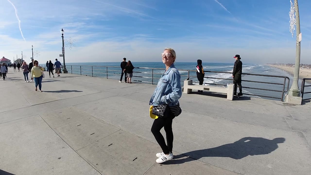 At 0:02, the woman is farther across the frame on the Huntington Beach pier, with a man near the railing, a bench, the ocean horizon, and the sandy shoreline visible behind her.