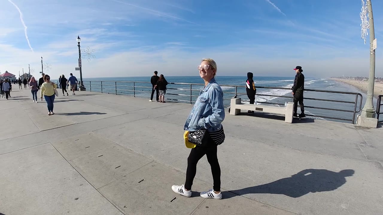 Woman walking on pier with bright sky, ocean, and scattered pedestrians.