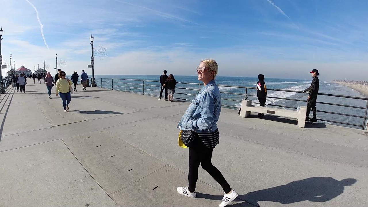 A woman in sunglasses and a denim jacket walks along a Huntington Beach pier while pedestrians, railings, surf, and a blue sky fill the background.