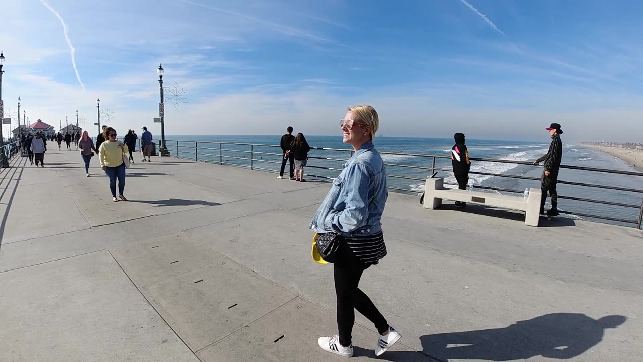 The walking woman is positioned near the right edge of the Huntington Beach pier frame as other visitors move along the walkway and surf breaks below.