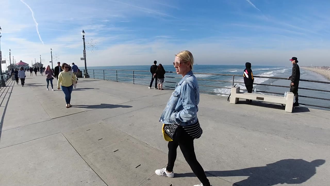 Foreground woman passes along a pier walkway as people stand near the ocean railing.