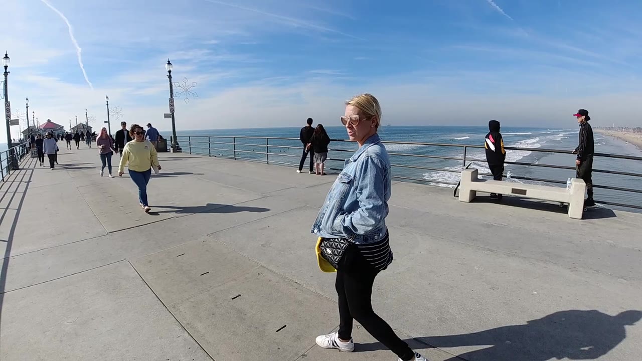 A woman moves near the right side of the frame on a Huntington Beach pier-style walkway while a person in yellow approaches and others stand by the railings over the surf.