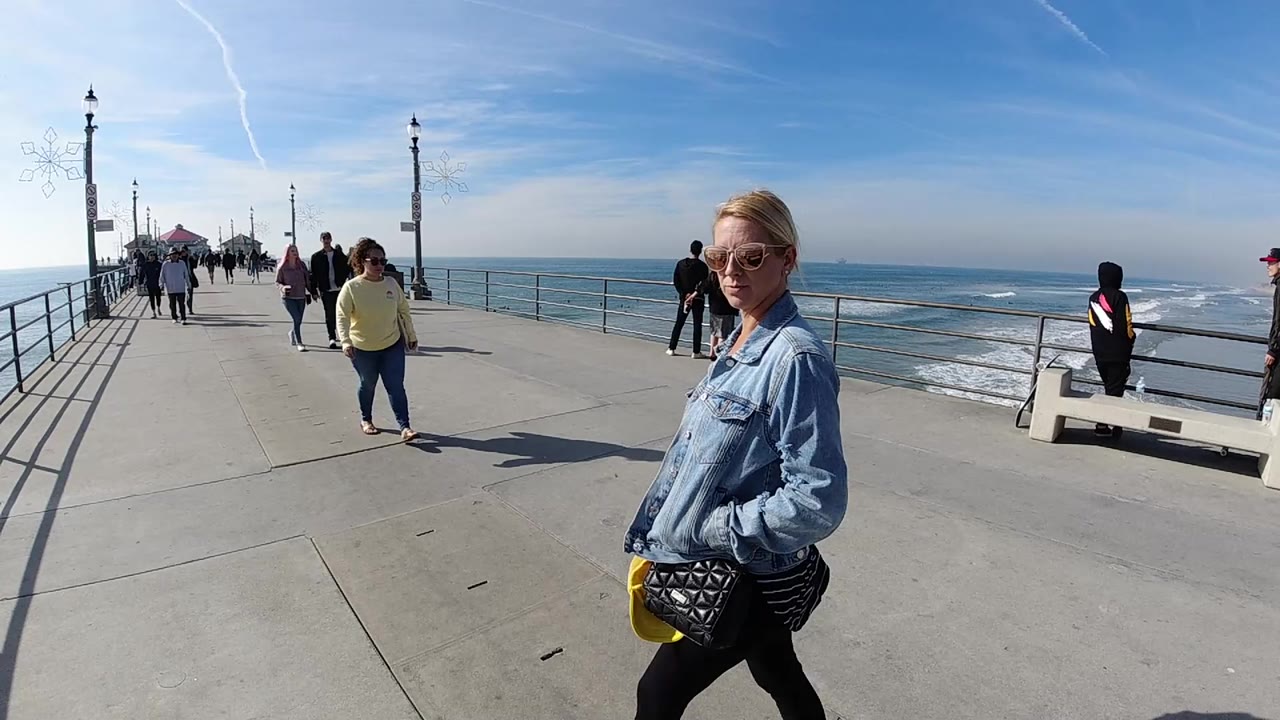 At Huntington Beach, California, a woman in sunglasses and a denim jacket walks close to the right side of frame while pedestrians move along the pier and waves roll beyond the railing.