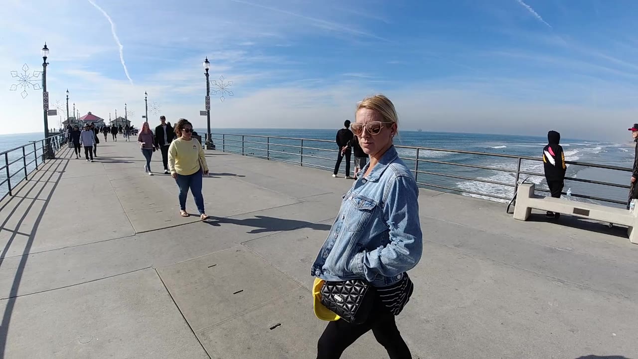 Sunny pier walkway with woman near camera, ocean railings, and people in the distance.