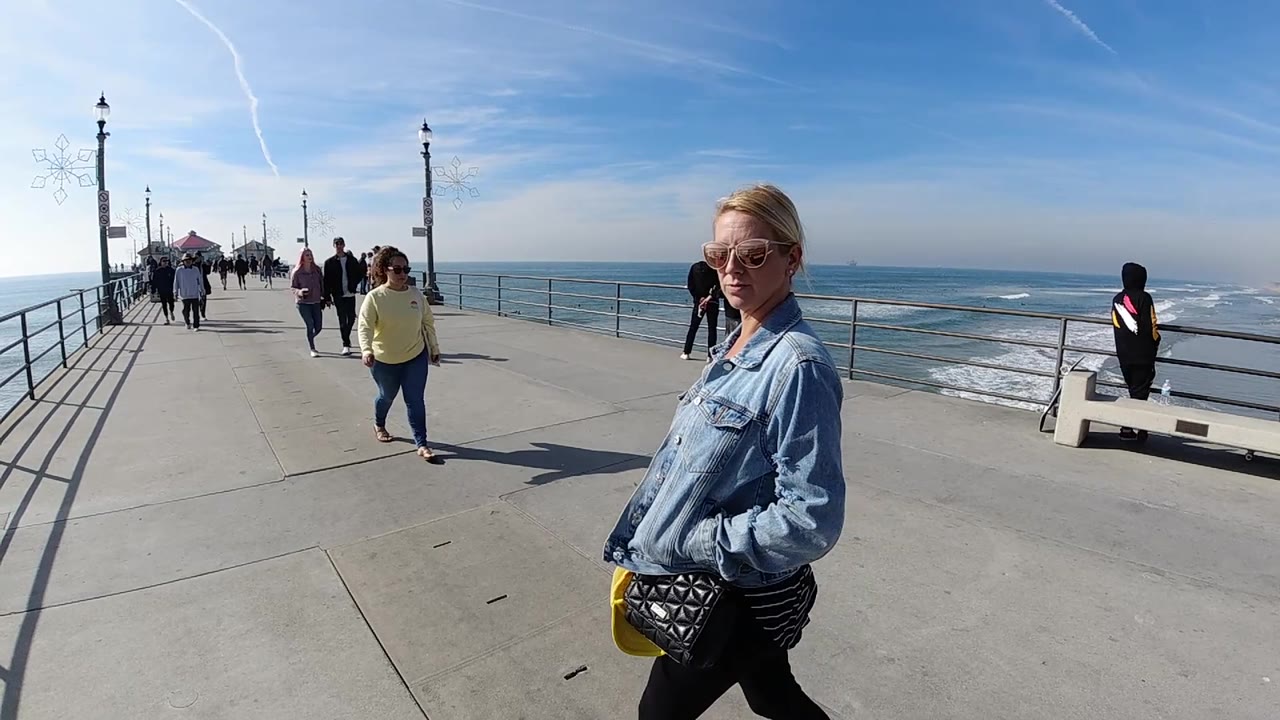 At Huntington Beach, California, the woman in denim is still close on the right while the pier opens ahead with scattered walkers, lamp posts, a bench area, and surf visible to the side.