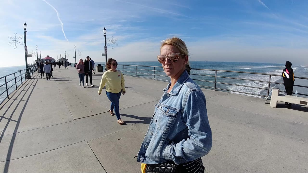 At 0:01, the foreground woman remains at right as a woman in a yellow top walks toward camera on the Huntington Beach pier, with surf and railings visible to the side.